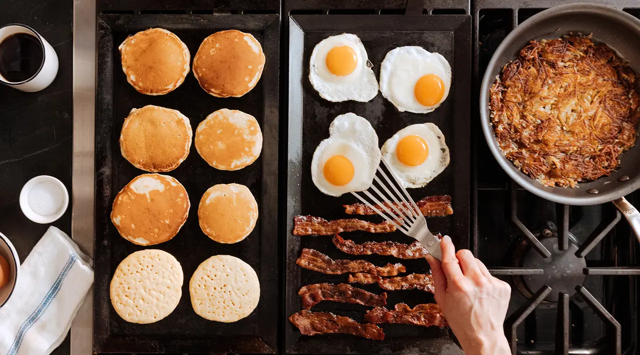 A variety of breakfast foods including pancakes, eggs, bacon, and hash browns are being cooked on a griddle and stove, with a person's hand flipping bacon.