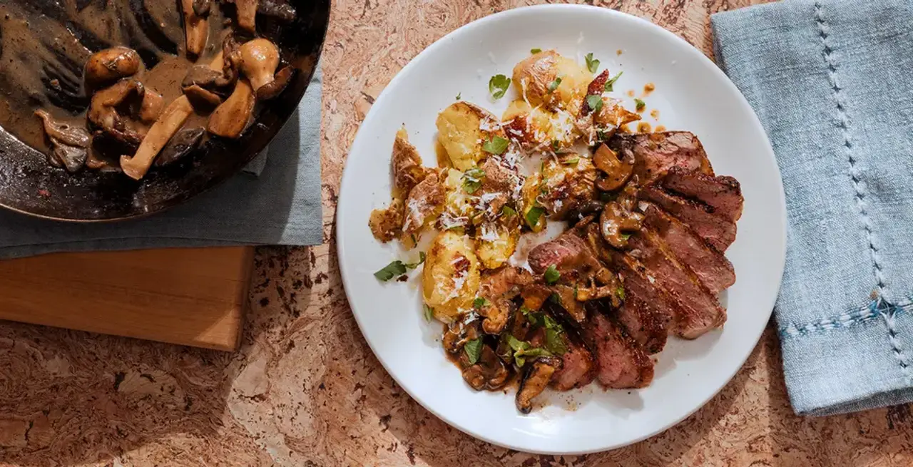 A well-plated meal consisting of steak and potatoes garnished with herbs and nuts, alongside a pan of sautéed mushrooms on a cork table surface.