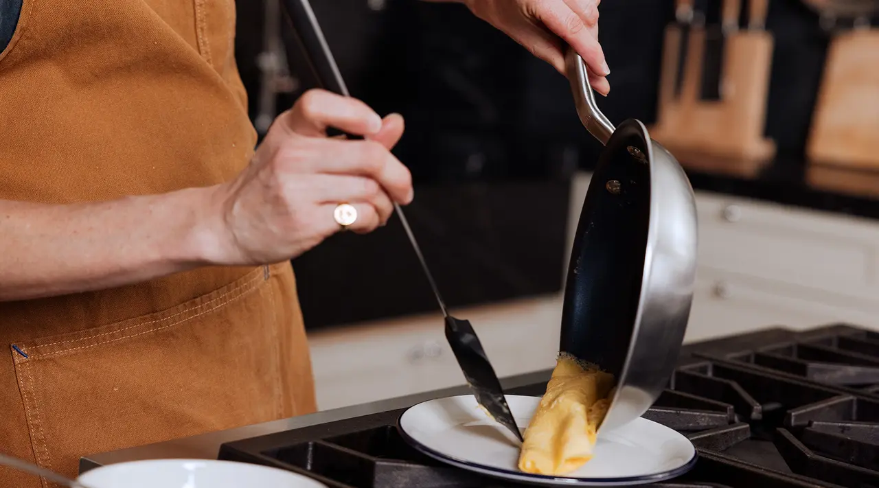 A person in an apron is carefully sliding an omelette from a pan onto a plate.