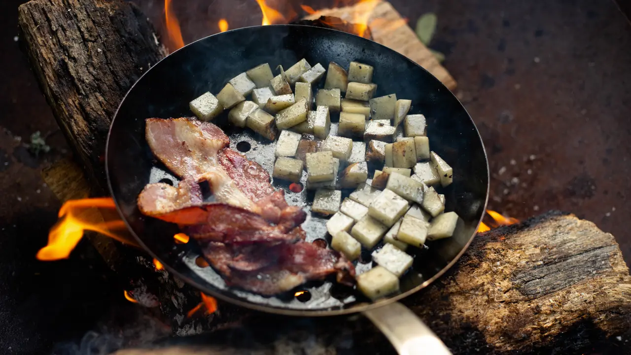 Bacon and diced food cook in a frying pan over an open campfire.