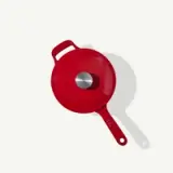 A red frying pan with a silver center and handle lies on a white background.