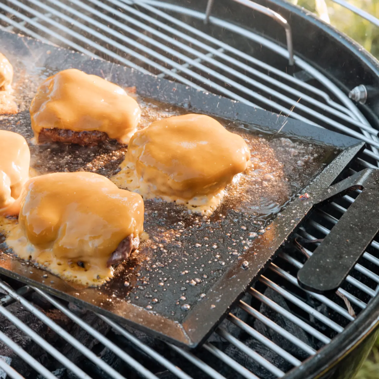 Juicy cheese-topped burgers sizzle on a flat grilling surface with a barbecue in the background.
