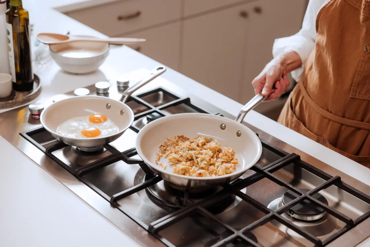 A person in an apron is cooking with two frying pans on a stove, with one pan containing eggs and the other sautéed ingredients.