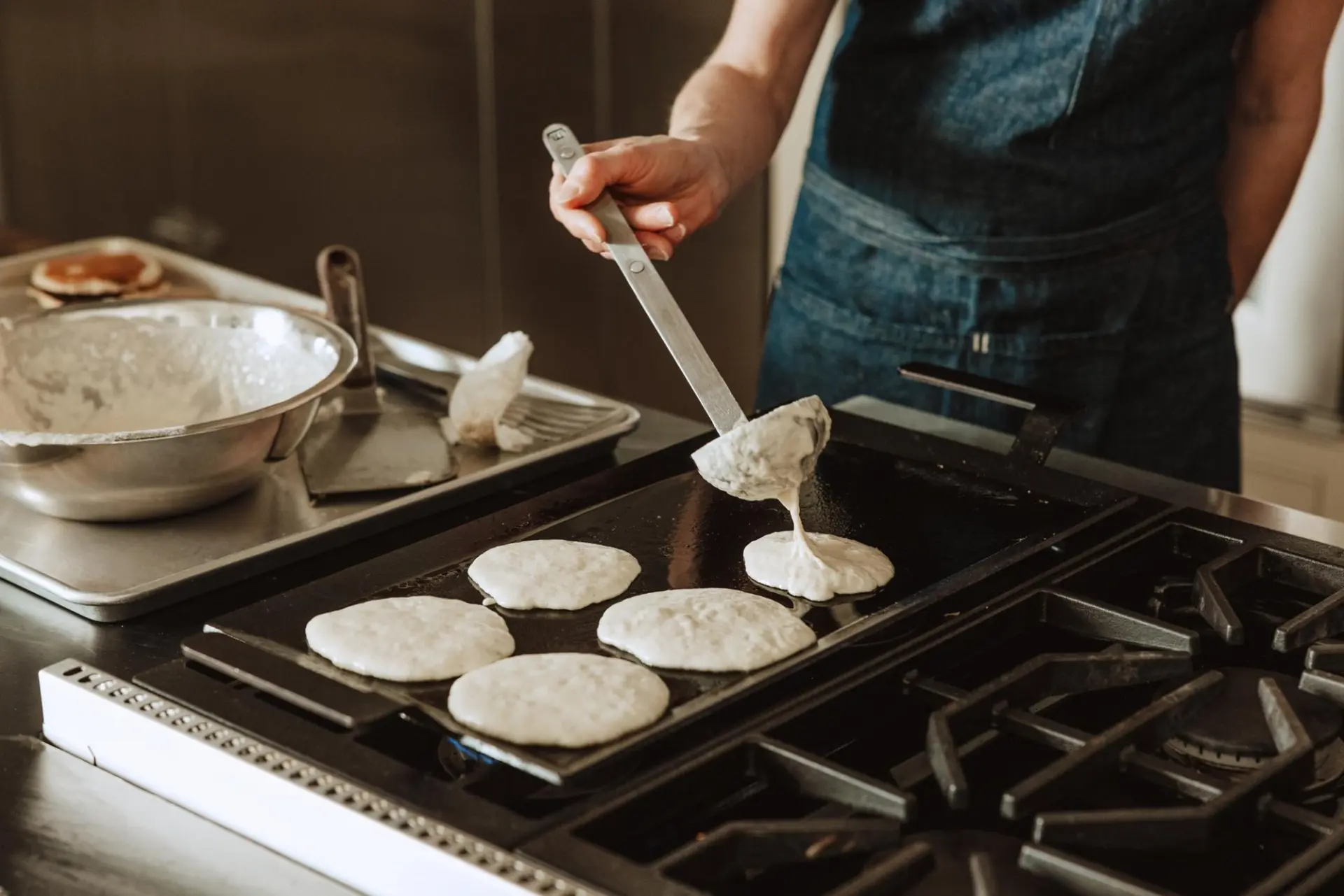 A person in an apron is pouring pancake batter onto a griddle, with additional ingredients visible in the background.
