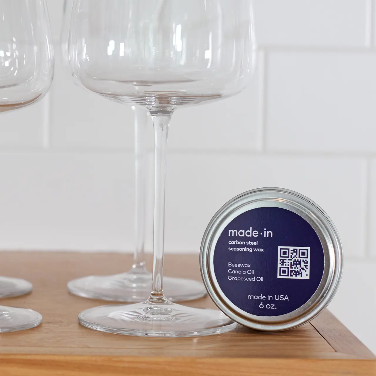 A tin of "made-in" brand carbon steel seasoning wax sits on a wooden surface in front of wine glasses, suggesting a kitchen setting.
