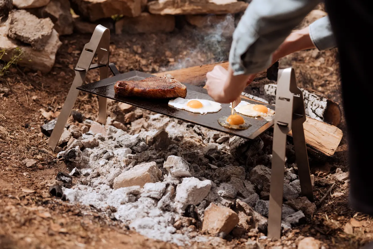 A person is cooking eggs and steak on a grill over an open fire outdoors.