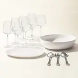 A collection of dining ware including plates, bowls, an assortment of glasses, and silverware is arranged neatly against a light background.
