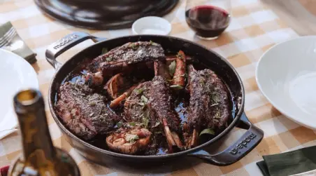 A skillet of grilled steaks is served on a checkered tablecloth next to a glass of red wine and dining ware.