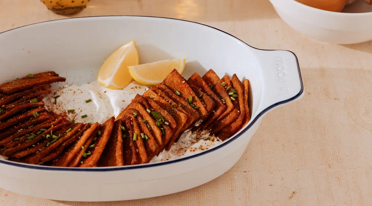 A dish of roasted carrots arranged in a fish-shaped pattern served with lemon wedges in a white oval ceramic dish, garnished with herbs on a kitchen countertop.