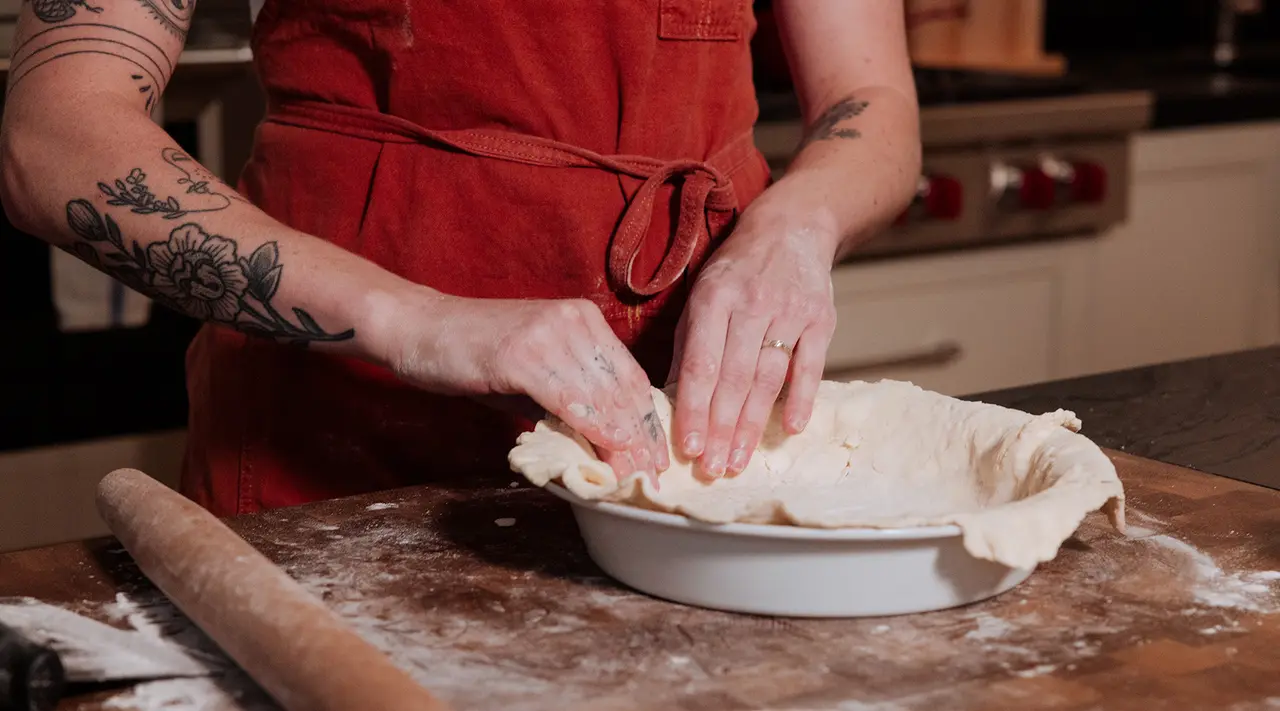 A person with tattooed arms is pressing pie dough into a dish on a flour-dusted surface with a rolling pin nearby.