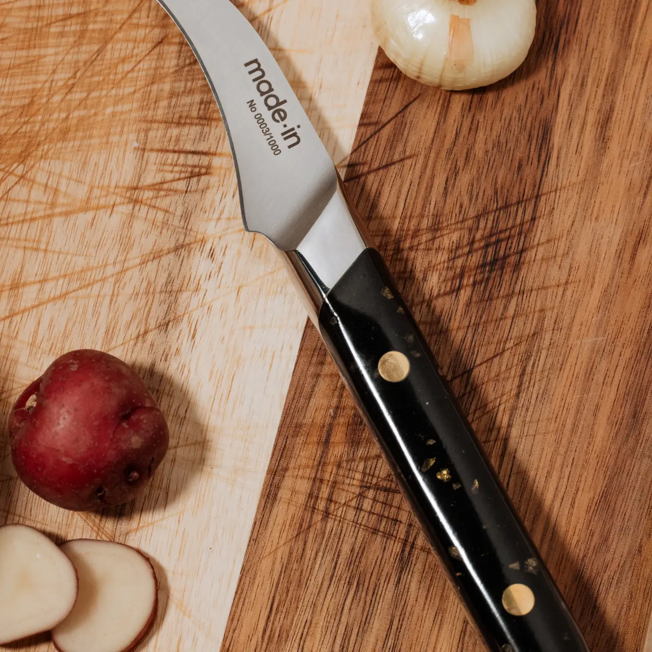 A chef's knife with a black handle is lying on a wooden cutting board next to a partially sliced apple and a whole onion.