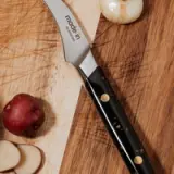 A chef's knife with a black handle is lying on a wooden cutting board next to a partially sliced apple and a whole onion.