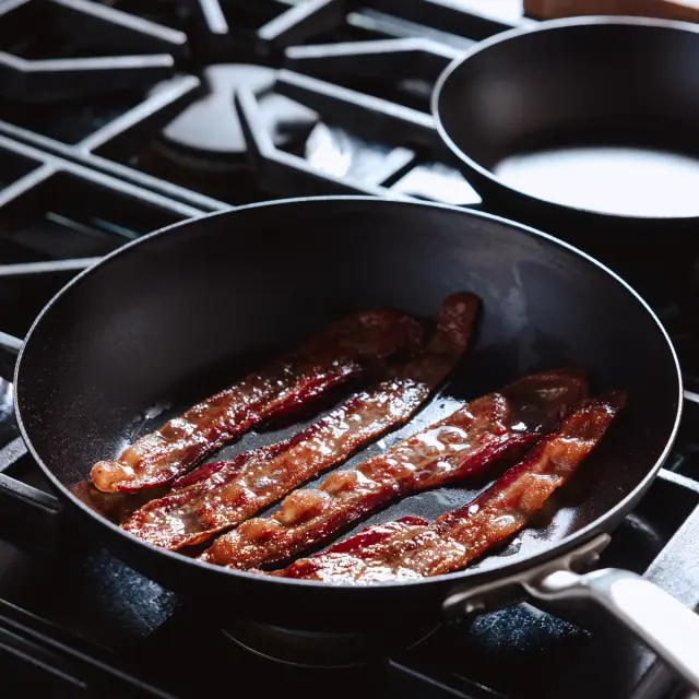 Sizzling bacon strips cooking in a black frying pan on a gas stove.