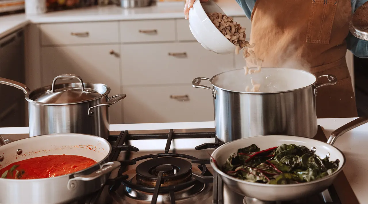 A person in an apron is pouring cooked meat into a pot on the stove, surrounded by pots of sauce and greens.