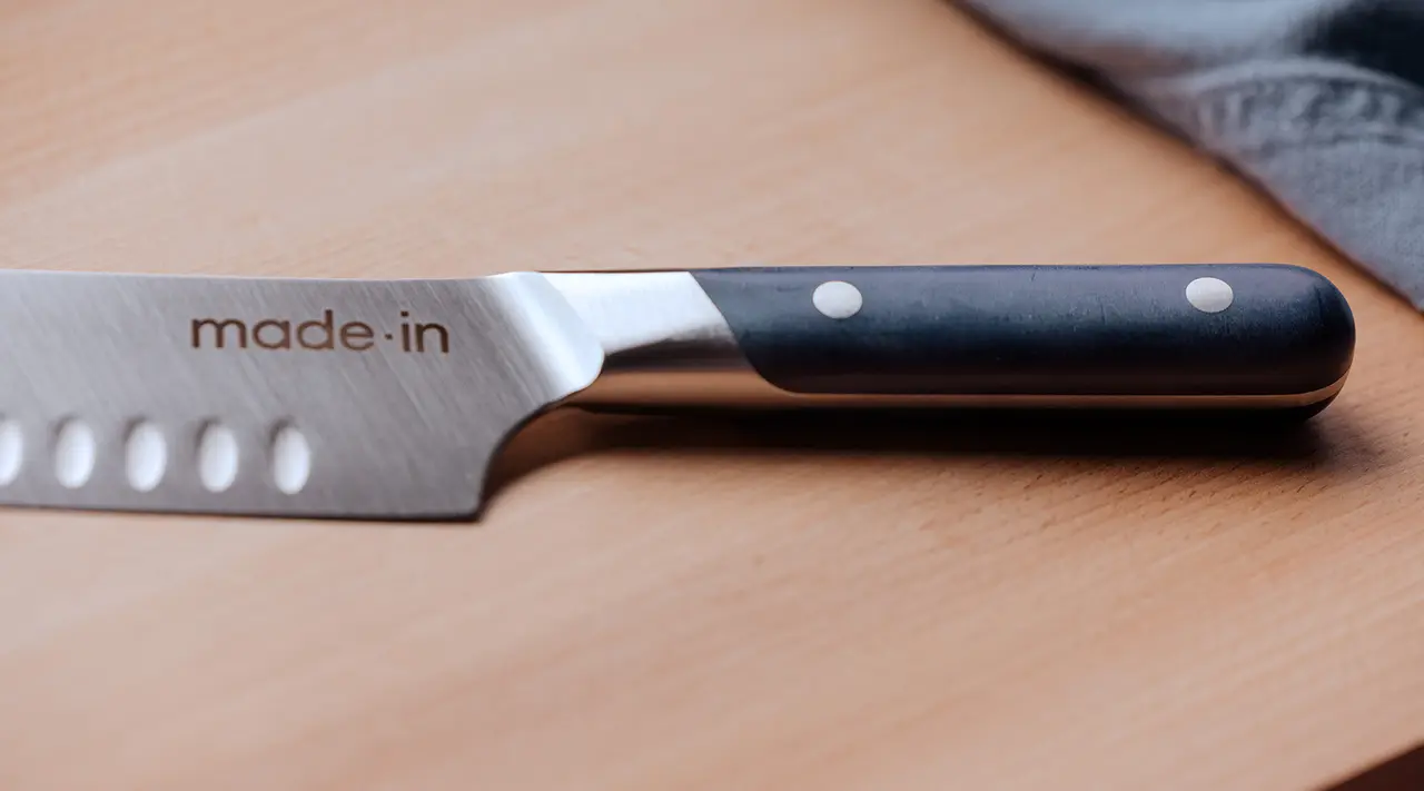 A close-up view of a stainless steel chef's knife with the words "made.in" on the blade and a black handle with three rivets, resting on a wooden surface next to a gray cloth.