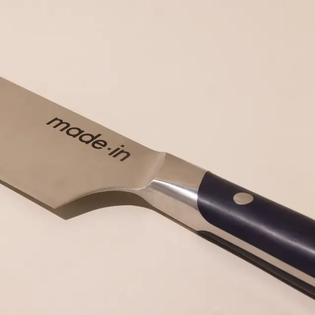 A close-up of a stainless steel kitchen knife with a blue and black handle, featuring the text "made.in" on the blade.