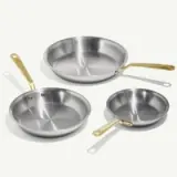 Three stainless steel frying pans with gold-colored handles are arranged in a staggered layout against a pale background.
