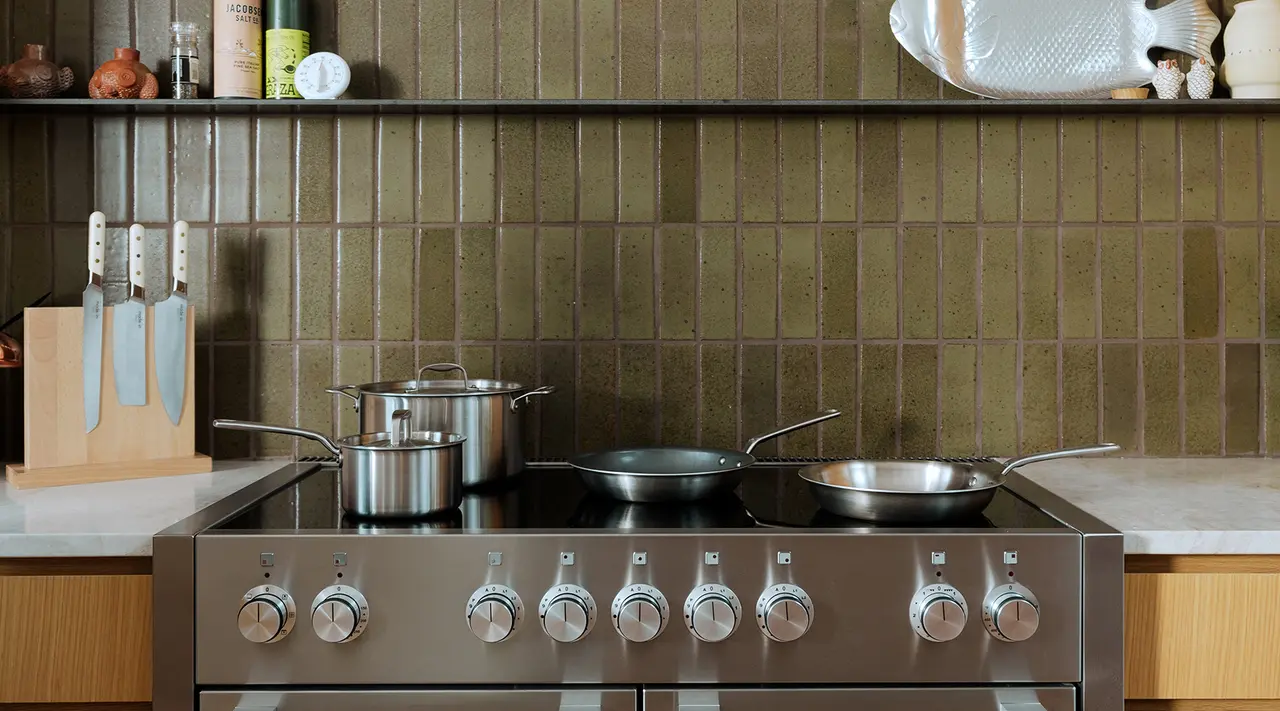 A stainless steel stovetop features a pot and two pans against a backdrop of olive green tiles, with kitchen utensils and spices arranged on the counter.