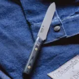A stainless steel knife with a black handle rests on a denim shirt featuring a branded tag and button.