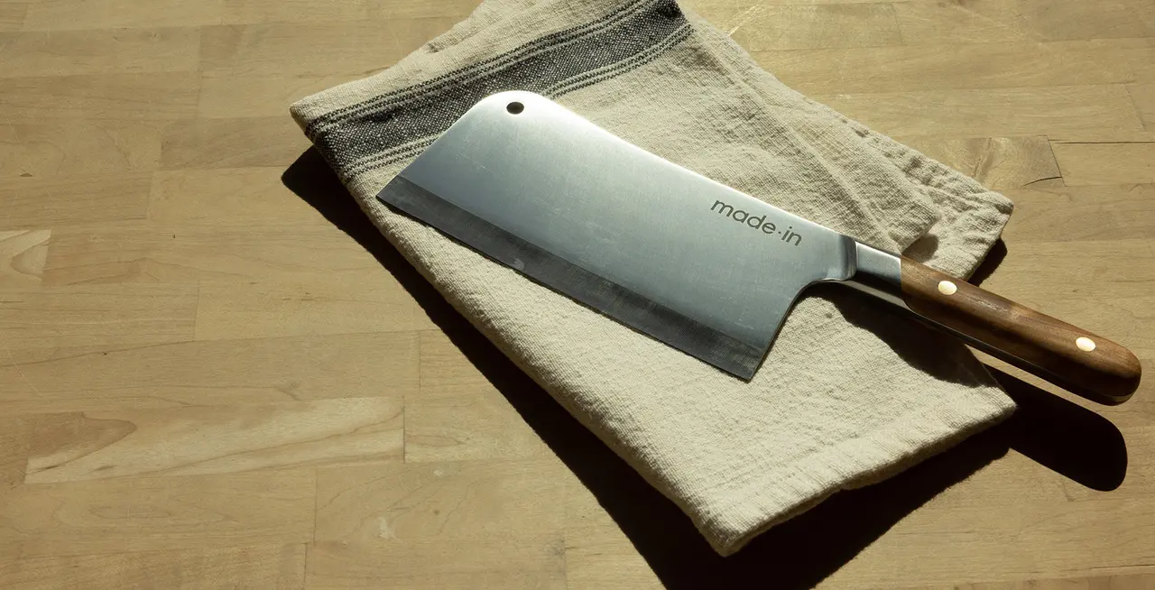A large kitchen knife with a wooden handle lies on a beige cloth on a wooden surface, casting a sharp shadow.