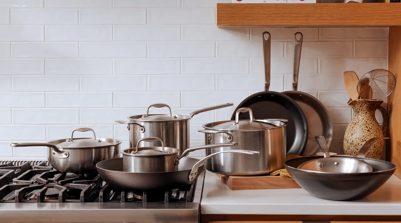 A variety of pots and pans are neatly arranged on a kitchen stovetop and counter against a white subway tile backsplash.