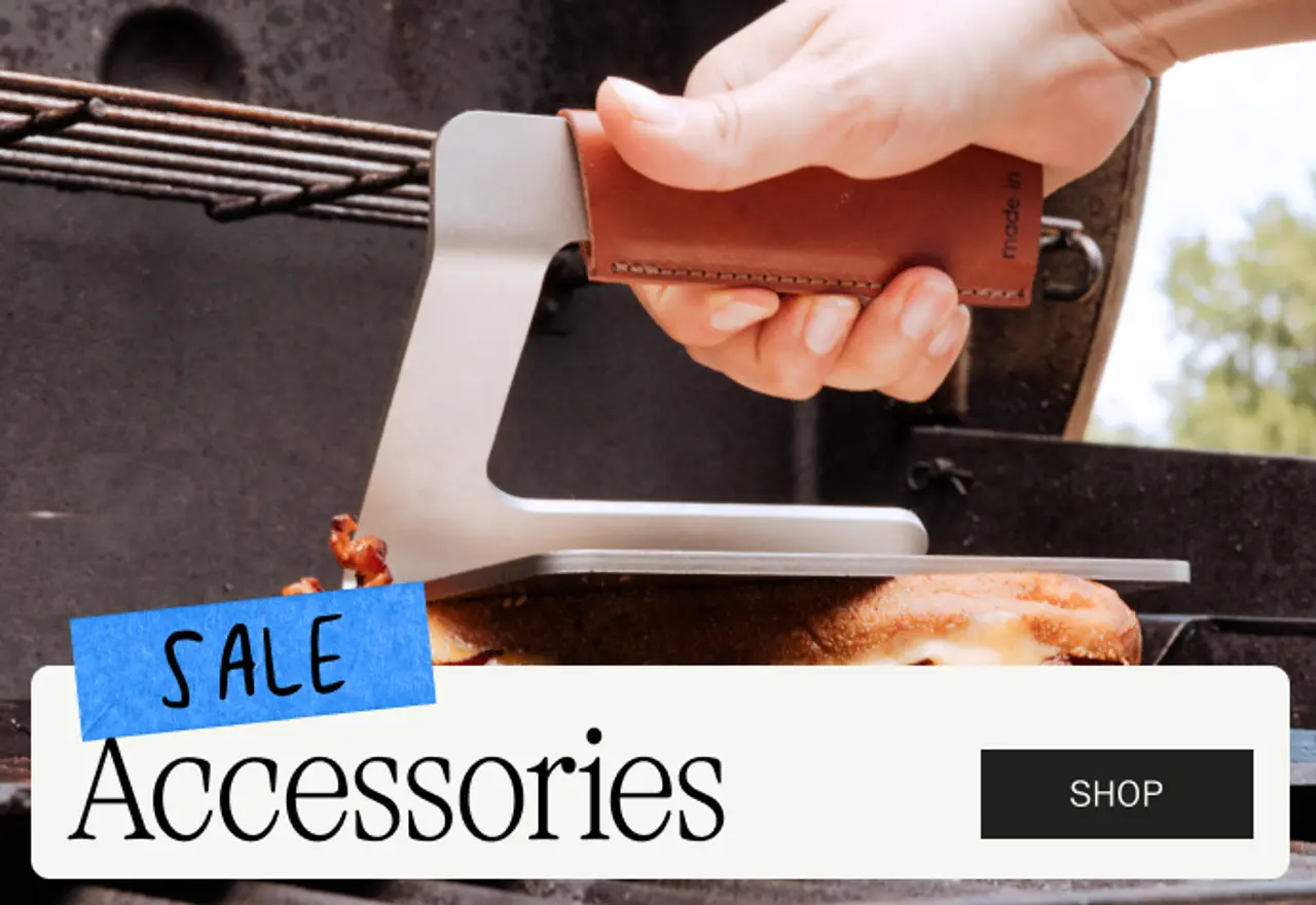 A hand grips a metallic spatula with a leather handle while cooking on a grill, accompanied by a text overlay promoting a sale on accessories.