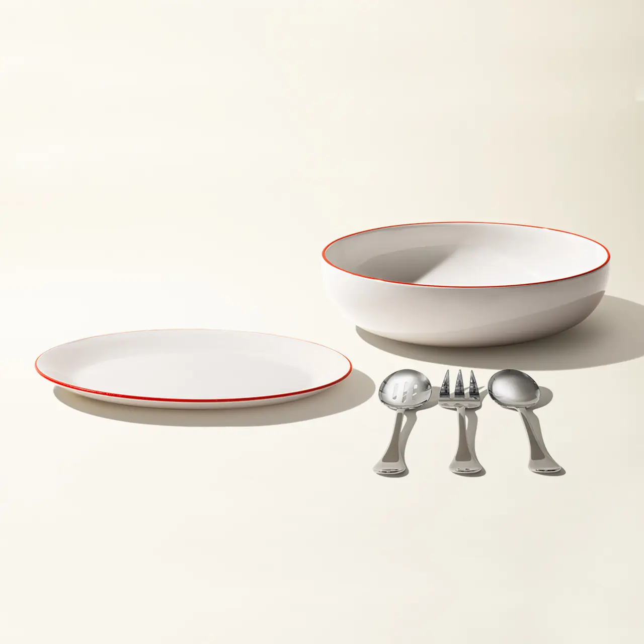 A set of simple white tableware with red trim, including a plate, a bowl, and four pieces of shiny silverware, is neatly arranged on a pale background.