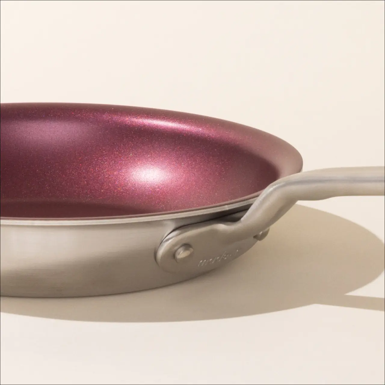 A close-up of a shiny purple non-stick frying pan with a stainless steel handle on a light background.