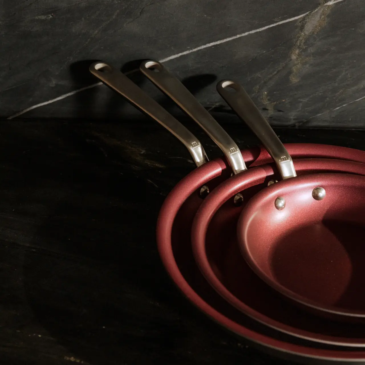 A set of three stacked red frying pans with stainless steel handles on a dark countertop.