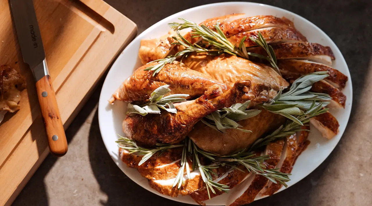 A roasted chicken garnished with rosemary sprigs is presented on a white plate, with sunlight casting shadows across the scene, accompanied by a carving knife and wooden board to the side.