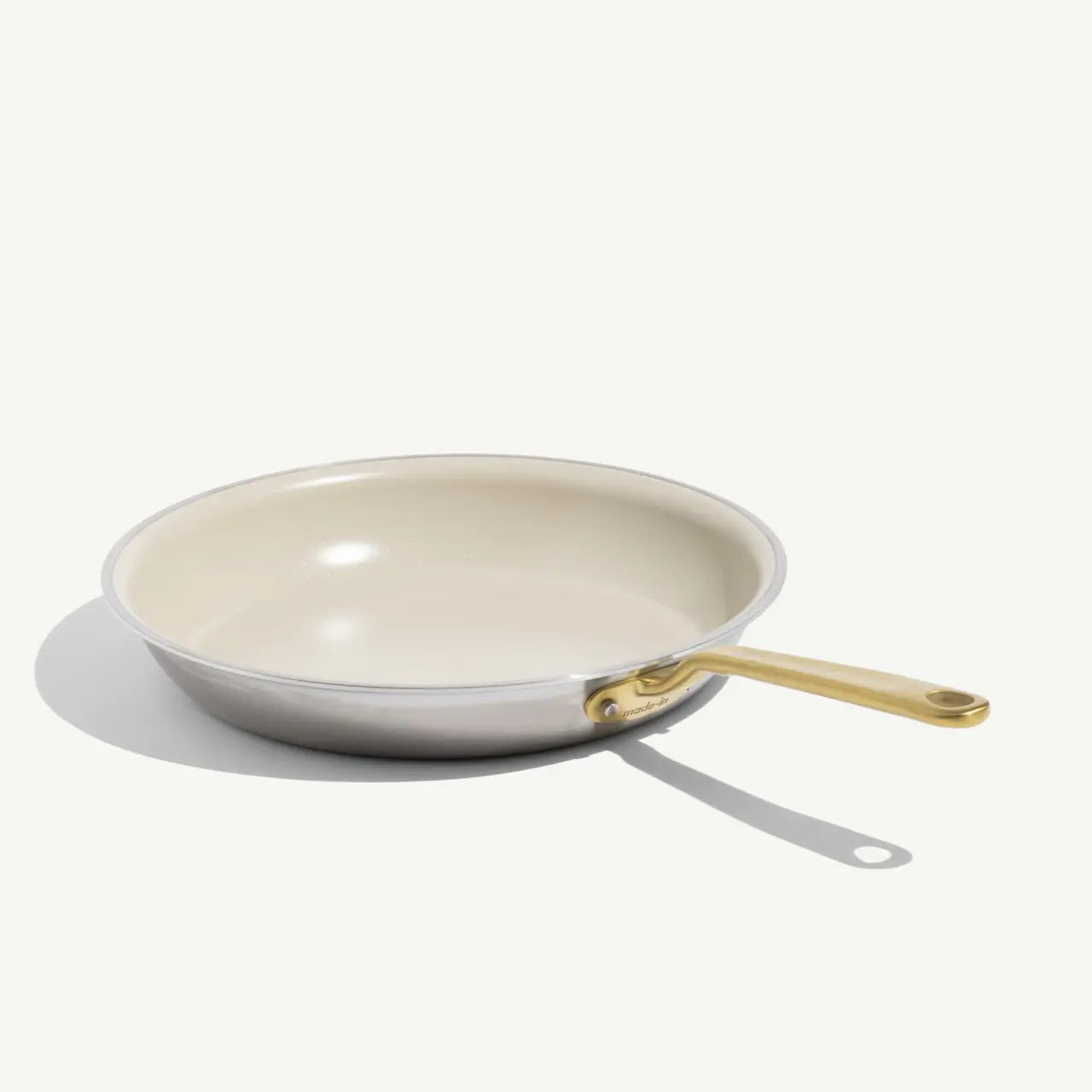 A white ceramic frying pan with a gold handle rests on a light background, casting a soft shadow.