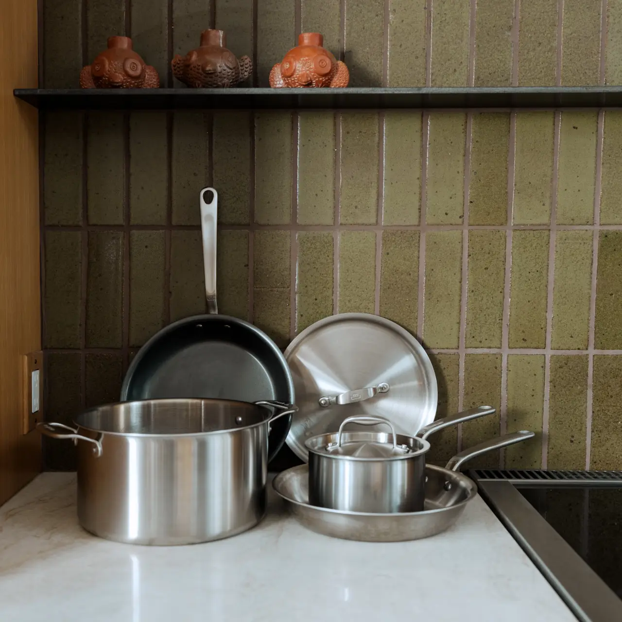 A neatly arranged set of stainless steel cookware sits on a kitchen counter beside a stovetop, with decorative items on the shelf above.