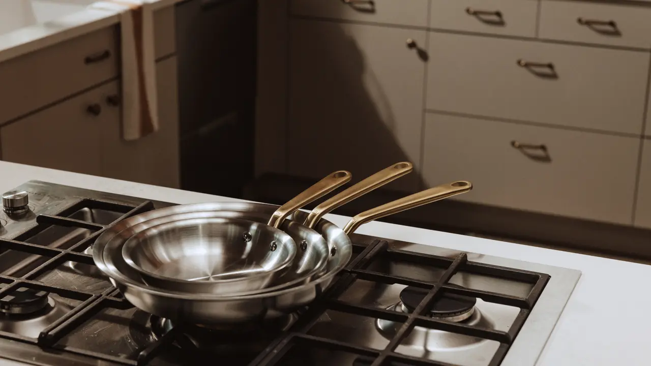 Stainless steel pans with golden handles sit on a gas stove in a modern kitchen.