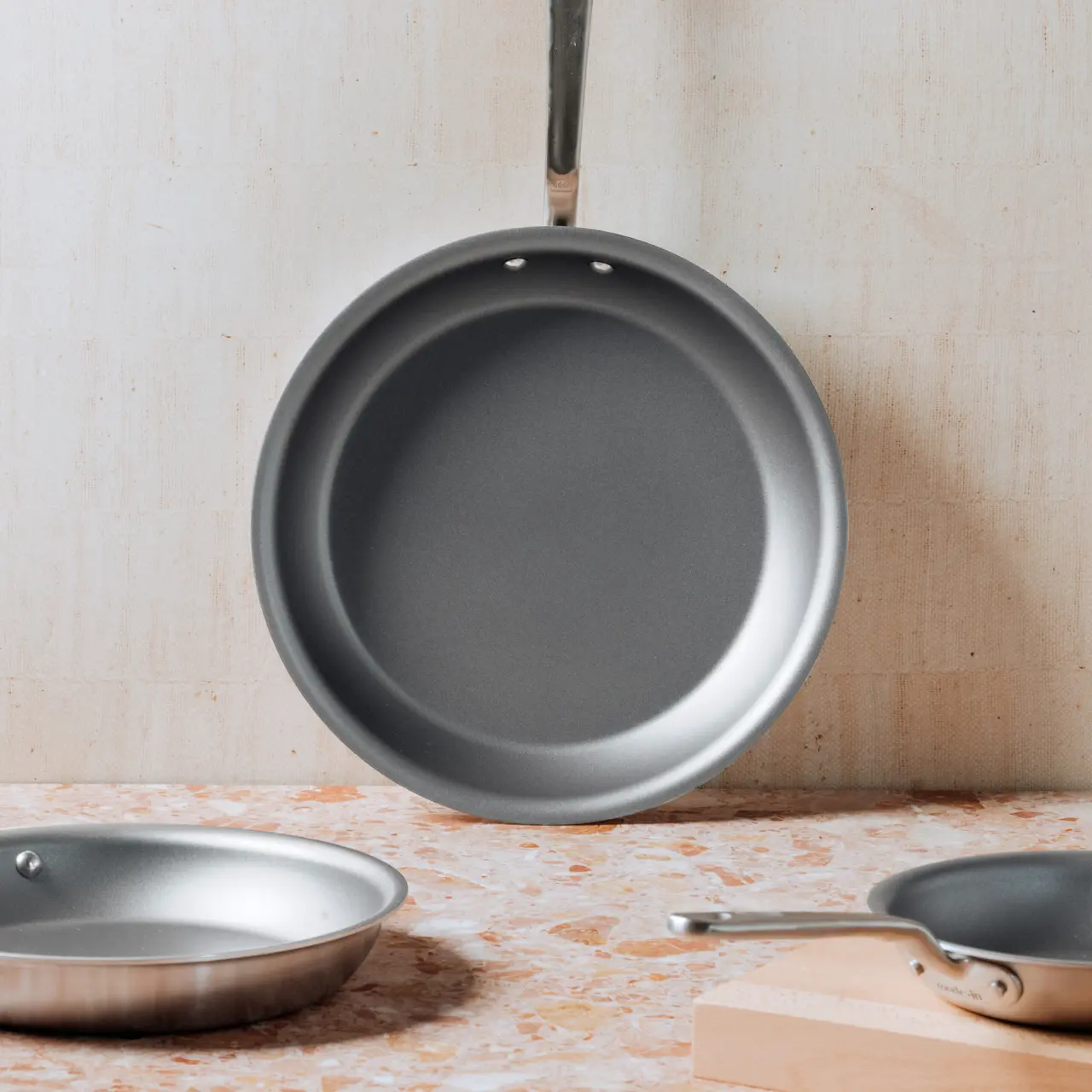 A non-stick frying pan hangs against a neutral backdrop with two other pans resting on a surface nearby.
