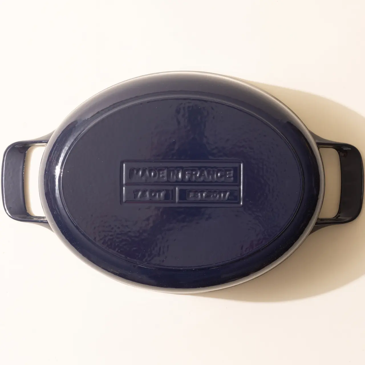 An oval, dark blue, enameled cast iron casserole dish with side handles sits against a light background.