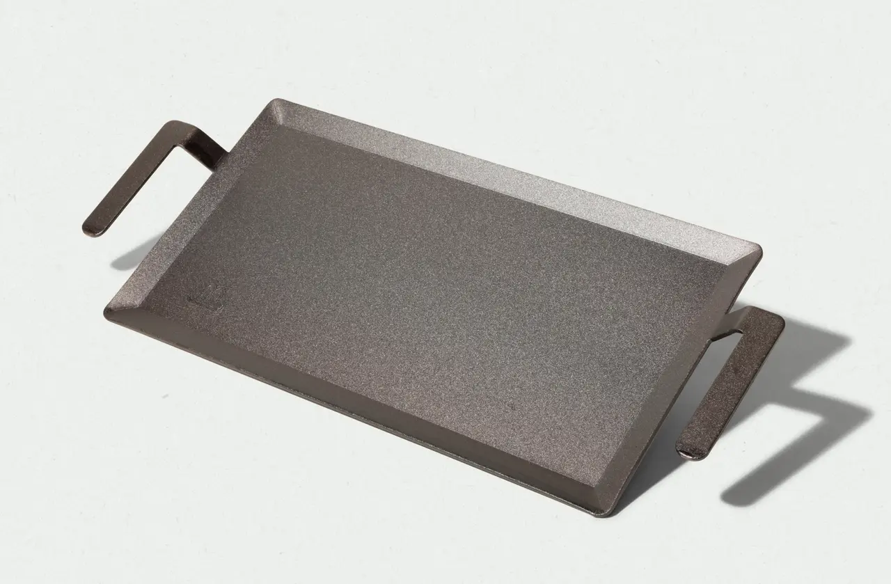 A cast iron griddle pan with handles rests on a light background, casting a soft shadow to its right.