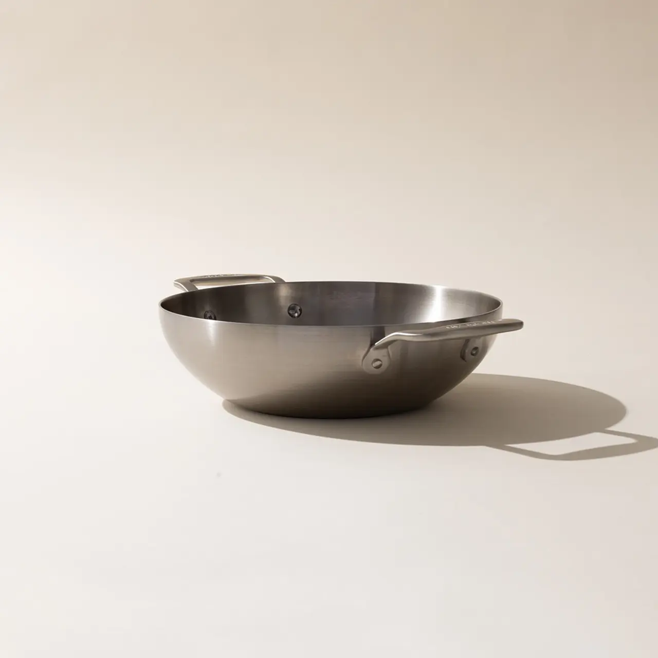 A stainless steel mixing bowl with riveted handles sits on a plain surface with its shadow visible.