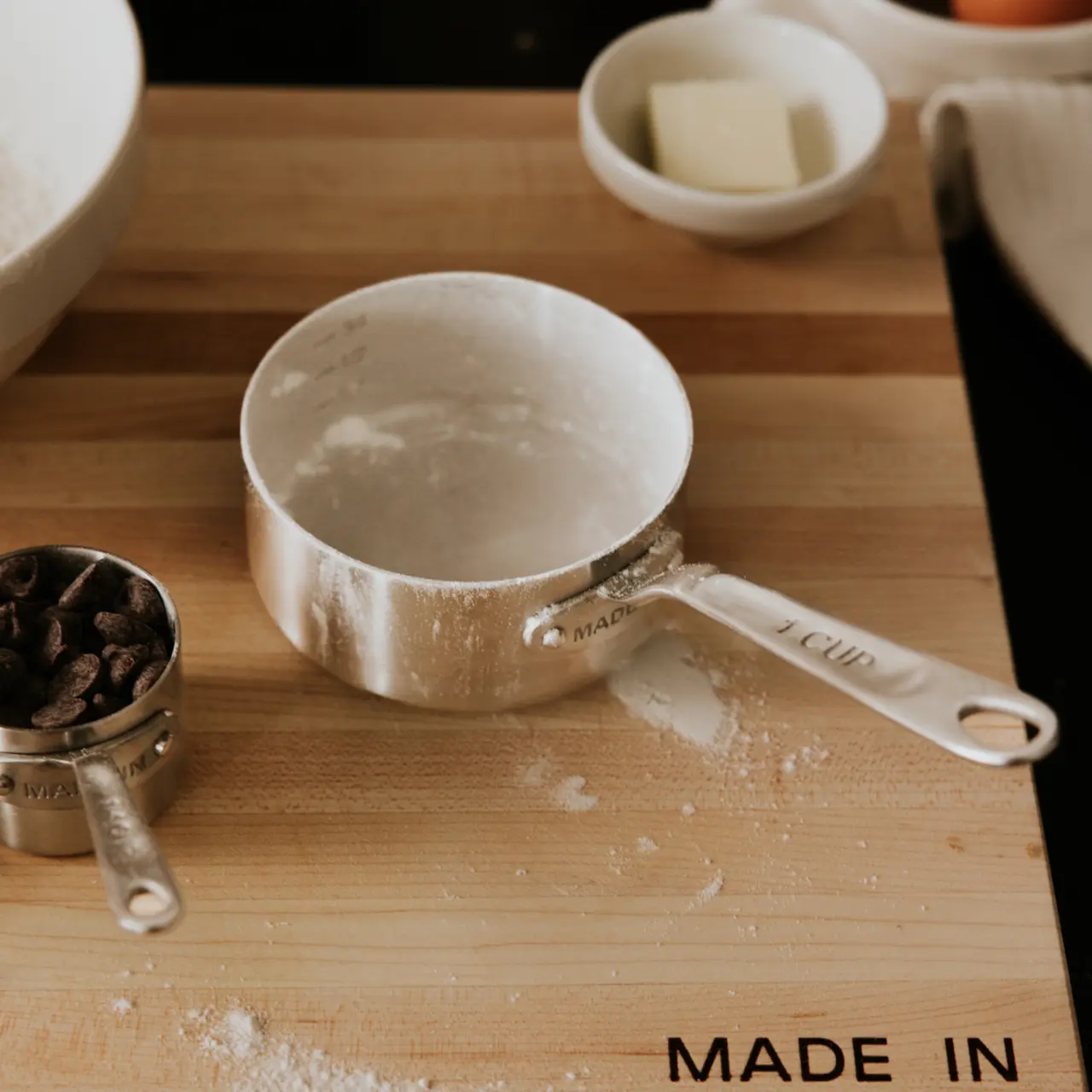 A dusting of flour covers a one-cup measuring scoop on a wooden cutting board amid various baking ingredients and utensils.