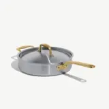 A round, silver cooking pot with a matching lid and gold handles sits on a light background.