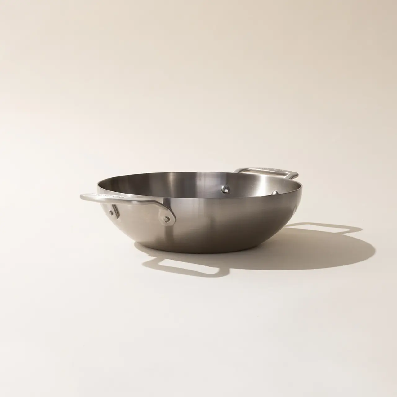 A stainless steel mixing bowl with handles on a neutral background casting a soft shadow.
