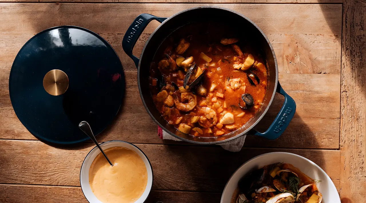 A hearty pot of seafood stew is accompanied by a bowl of sauce and some shellfish on a wooden table.