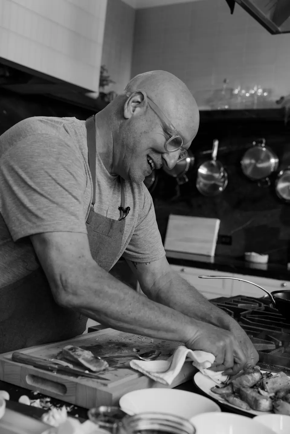 A black and white photo of a bald man in glasses smiling as he focuses on cooking in a kitchen filled with utensils and pots.