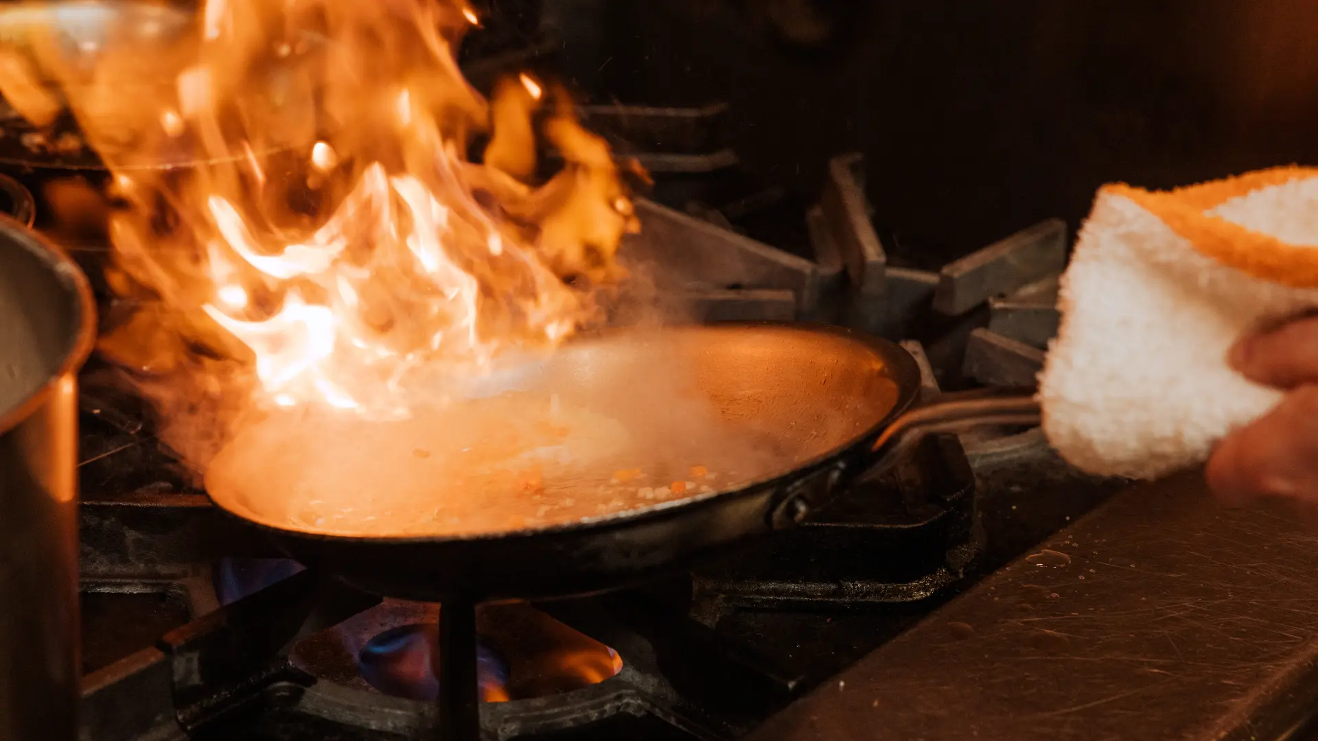 A pan on a stove is engulfed in flames while a person holds a towel nearby.