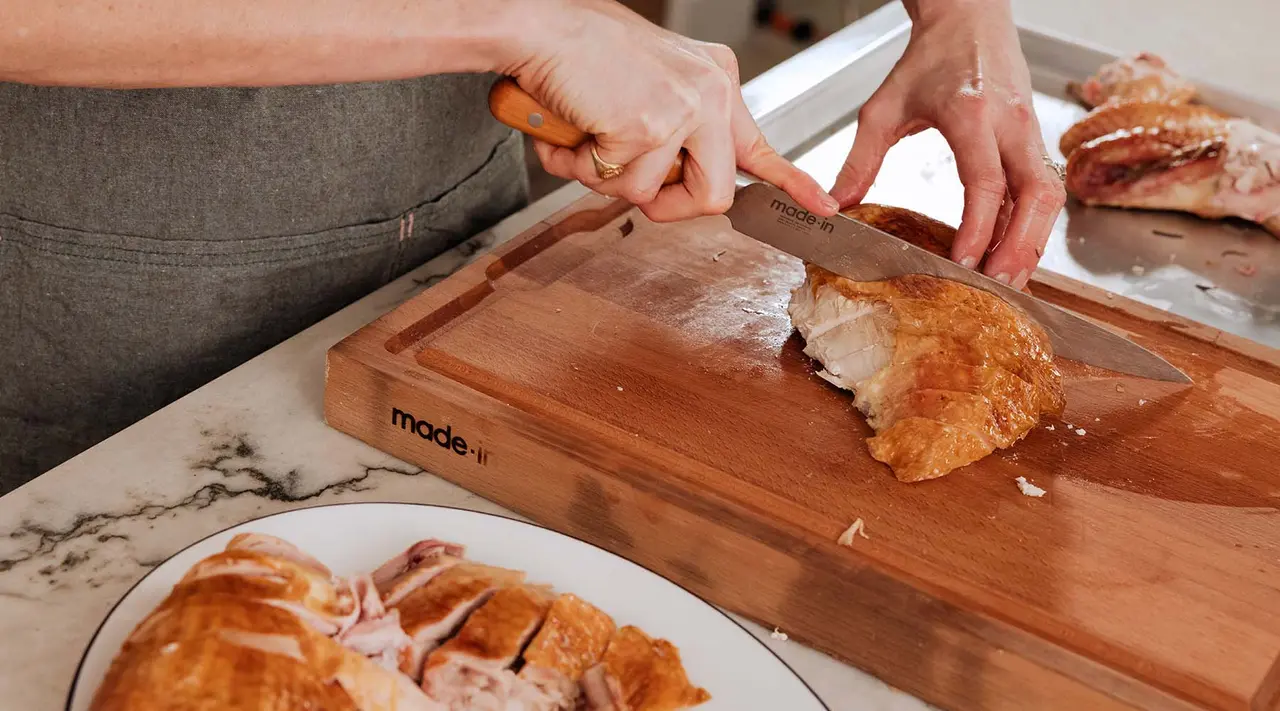 A person is slicing a crispy, golden-brown croissant on a wooden cutting board, with pieces of croissant already cut and placed on the plate nearby.
