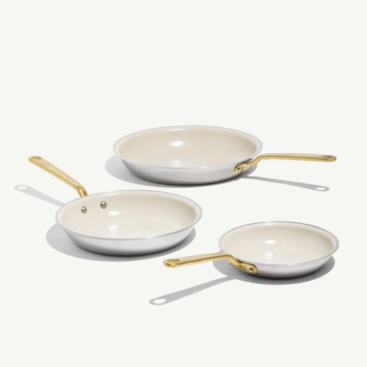 Three frying pans in varying sizes with sleek white interiors and gold handles are displayed against a light background.