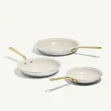 Three frying pans in varying sizes with sleek white interiors and gold handles are displayed against a light background.