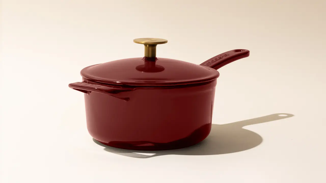 A red saucepan with a matching lid and a gold-tone handle knob on a neutral background with a soft shadow.