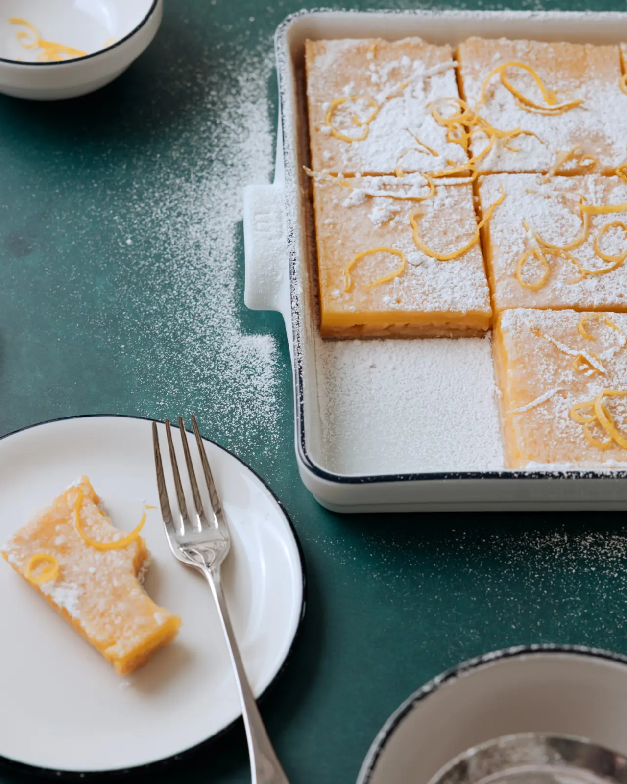 A baking dish holds lemon bars dusted with powdered sugar on a green surface, with one bar served on a plate beside it.