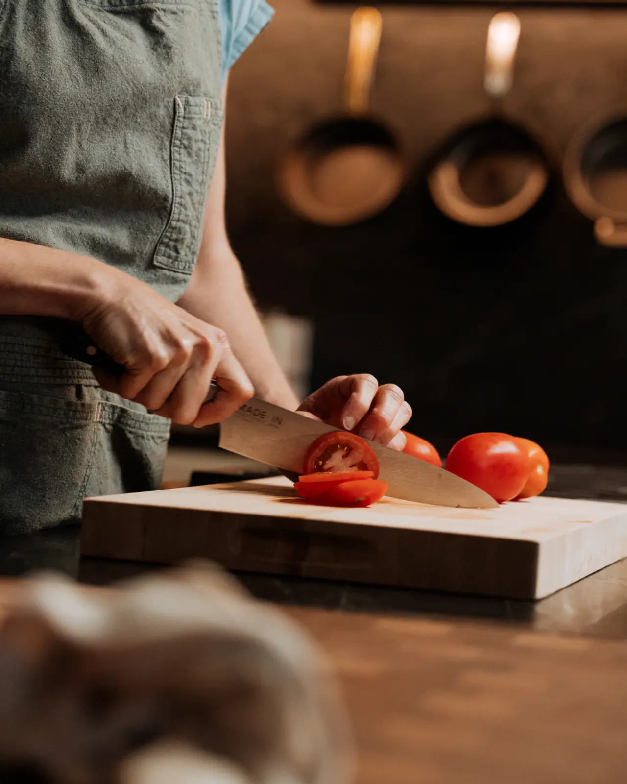 A person wearing an apron slices tomatoes on a wooden cutting board in a kitchen.