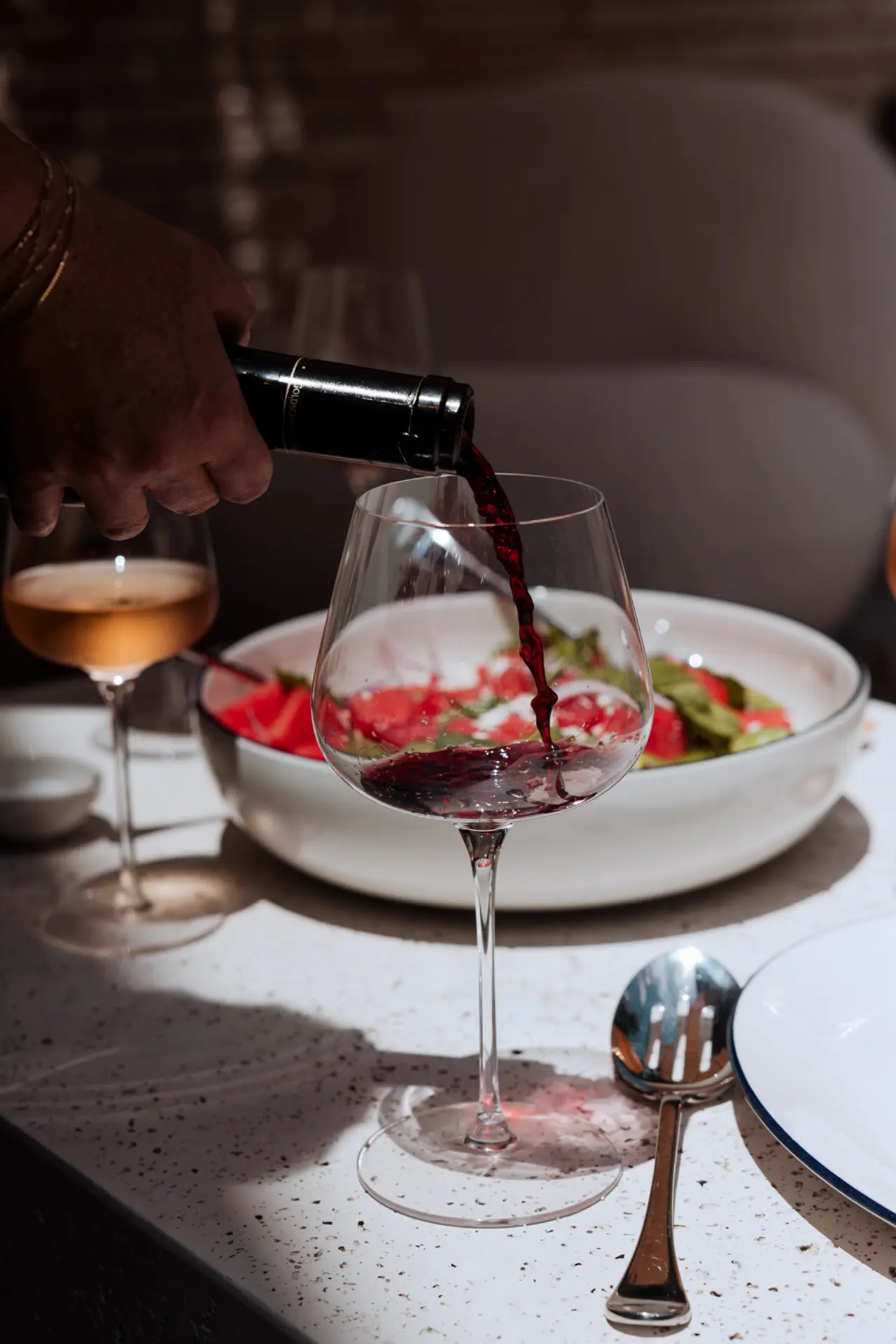 Red wine is being poured into a stemmed glass at a dining table with plates of food and another glass of beverage.